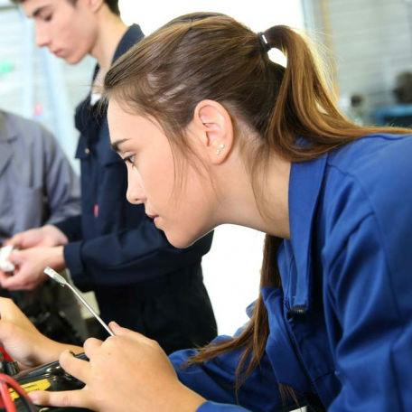 Image shows a female apprentice in blue overalls, holding a tool whilst leaning over a piece of equipment
